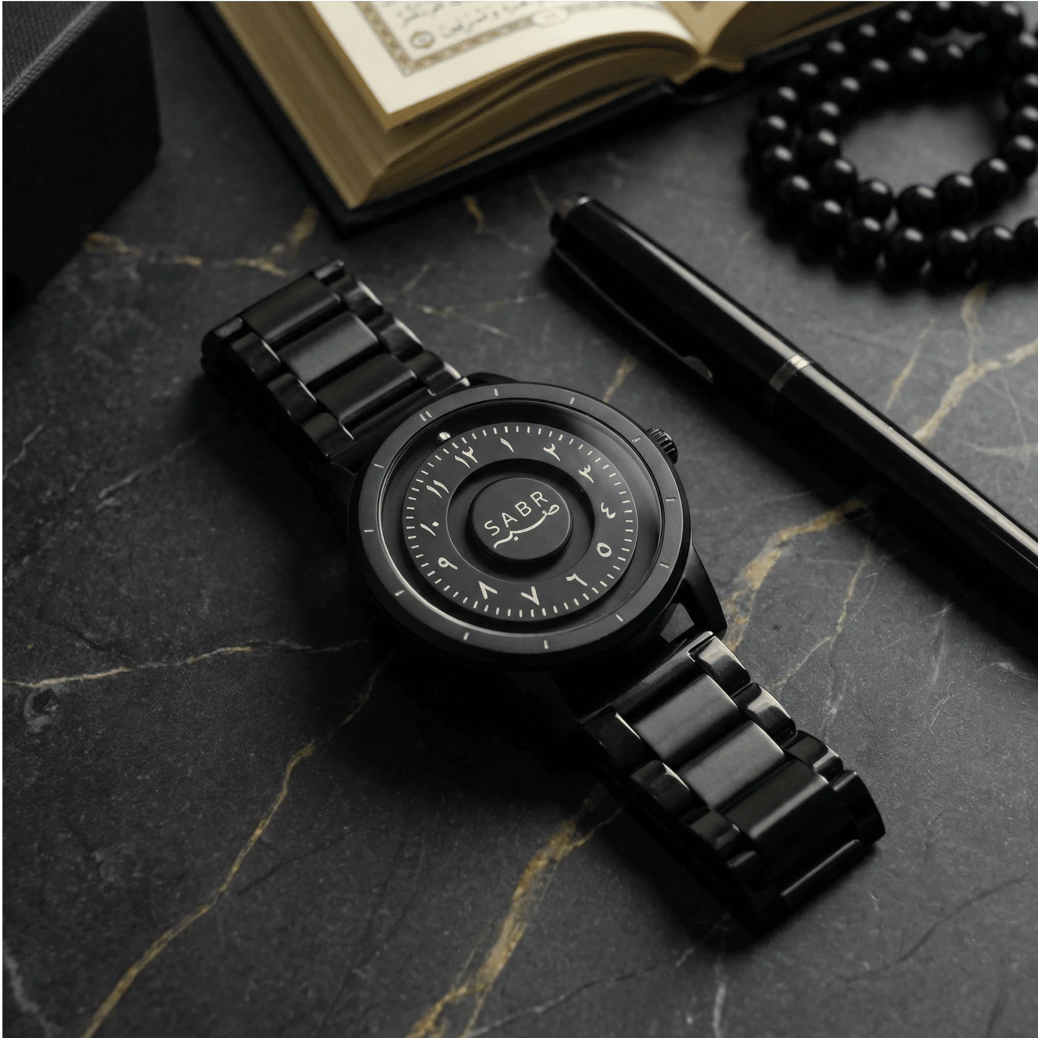 Black sabr watch on a dark marble surface with a book and beads in the background